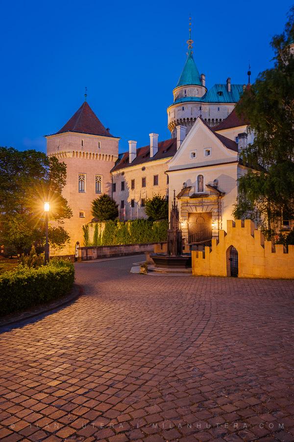 Bojnice Castle with illuminated towers and a cobblestone path during blue hour.