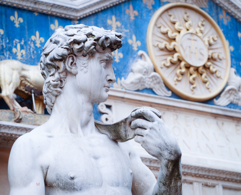 Close-up photograph of the David statue replica in front of the ornate blue and gold facade of Palazzo Vecchio in Florence, Italy