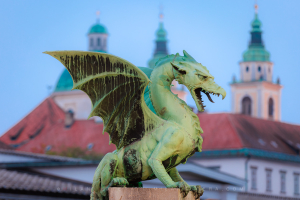 Dragon Bridge at dawn in Ljubljana, Slovenia, featuring a close-up of the iconic green dragon statue with the blurred St. Nicholas Cathedral in the background.