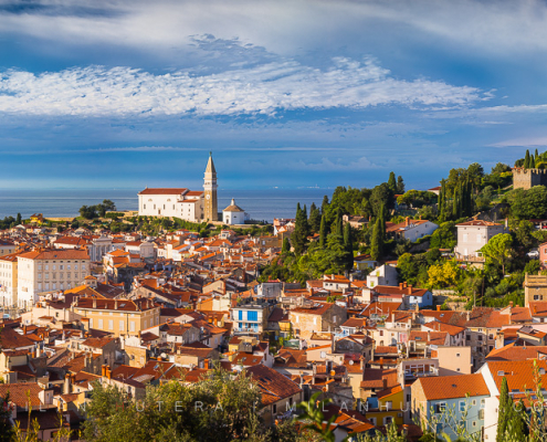 Panorama of Piran, Slovenia, featuring red-roofed buildings, the St. George's Parish Church bell tower, and medieval walls on a hillside, overlooking a marina and the blue Adriatic Sea