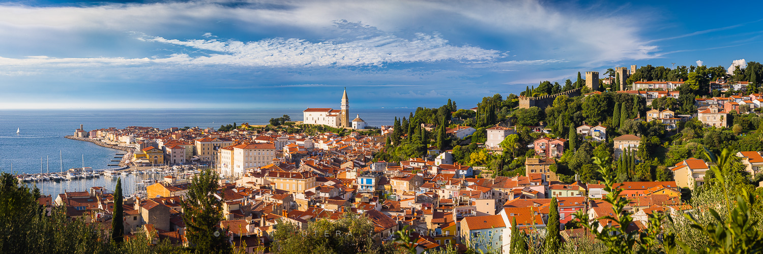 Panorama of Piran, Slovenia, featuring red-roofed buildings, the St. George's Parish Church bell tower, and medieval walls on a hillside, overlooking a marina and the blue Adriatic Sea