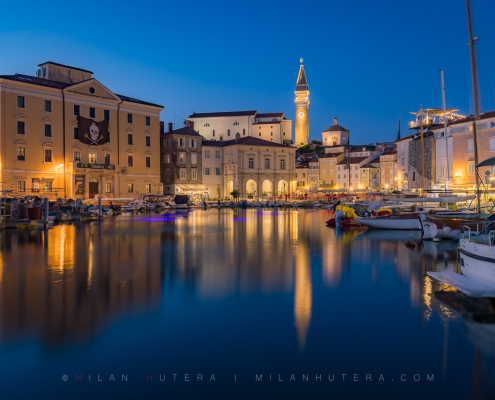 Twilight photo of Piran harbor featuring St. George's Parish Church bell tower reflections and a pirate flag from the historic Pomorski Krst festival, Slovenia.