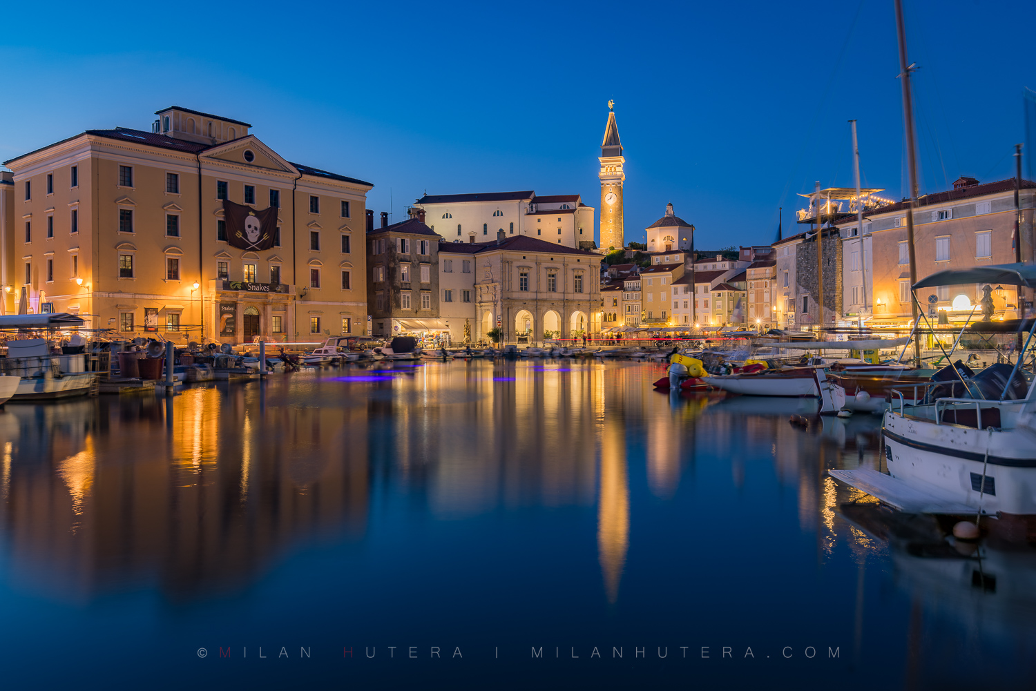 Twilight photo of Piran harbor featuring St. George's Parish Church bell tower reflections and a pirate flag from the historic Pomorski Krst festival, Slovenia.