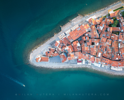Top-down aerial drone panorama of the Piran peninsula at sunrise, showcasing the dense red-tiled roofs caught in the first morning light, the historic lighthouse church, and a small boat leaving a wake in the calm, turquoise Adriatic Sea.