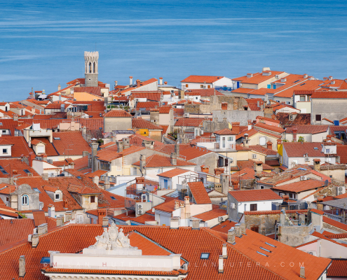 Piran rooftops view with a sea of orange terracotta tiles, the Church of Our Lady of Health lighthouse, and a smooth Adriatic Sea.