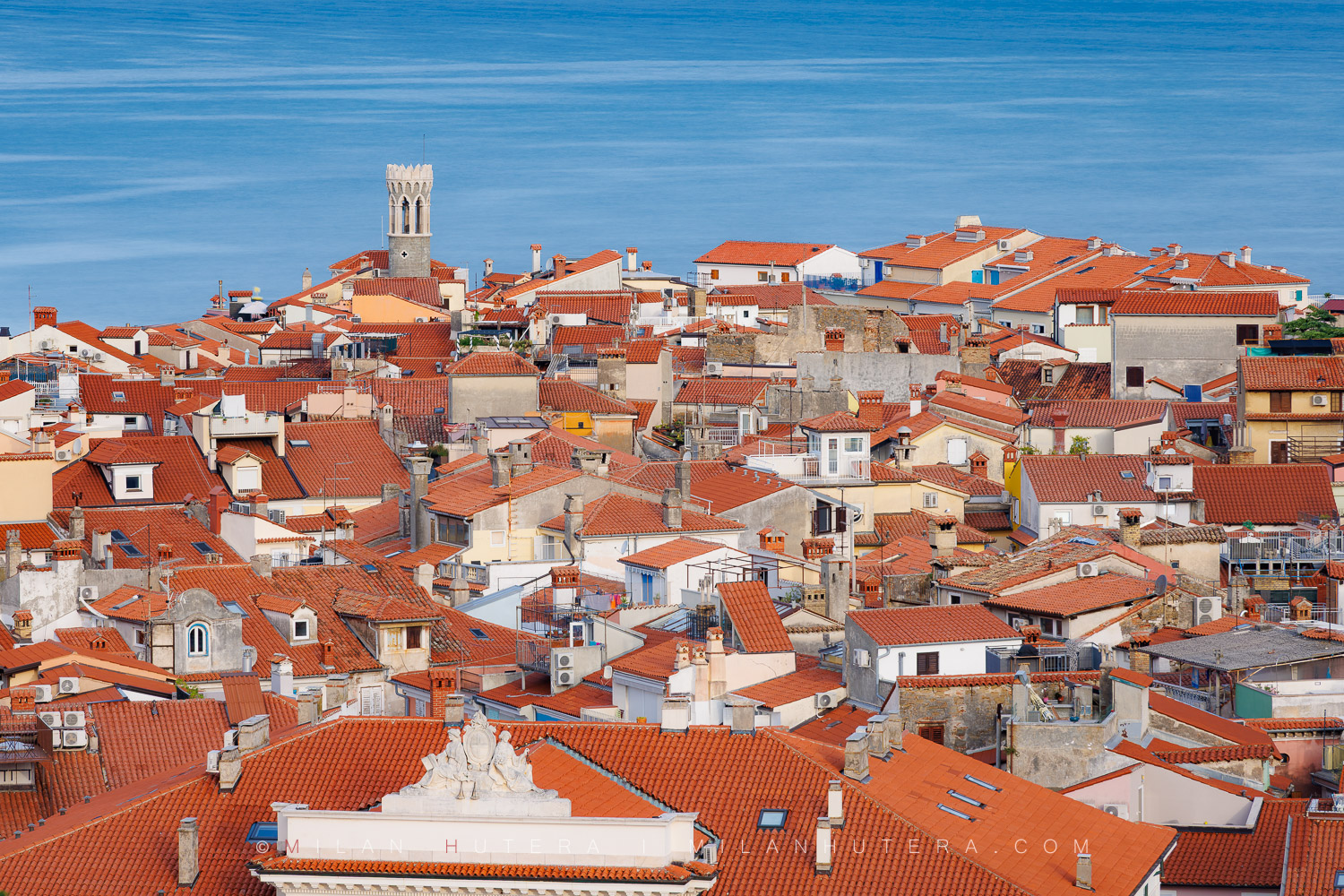 Piran rooftops view with a sea of orange terracotta tiles, the Church of Our Lady of Health lighthouse, and a smooth Adriatic Sea.