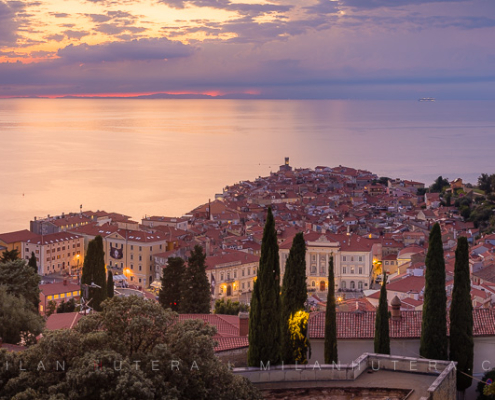 Panoramic sunset view of Piran old town from the Piran Walls, Slovenia, featuring the St. George's Parish Church bell tower and the Adriatic Sea.