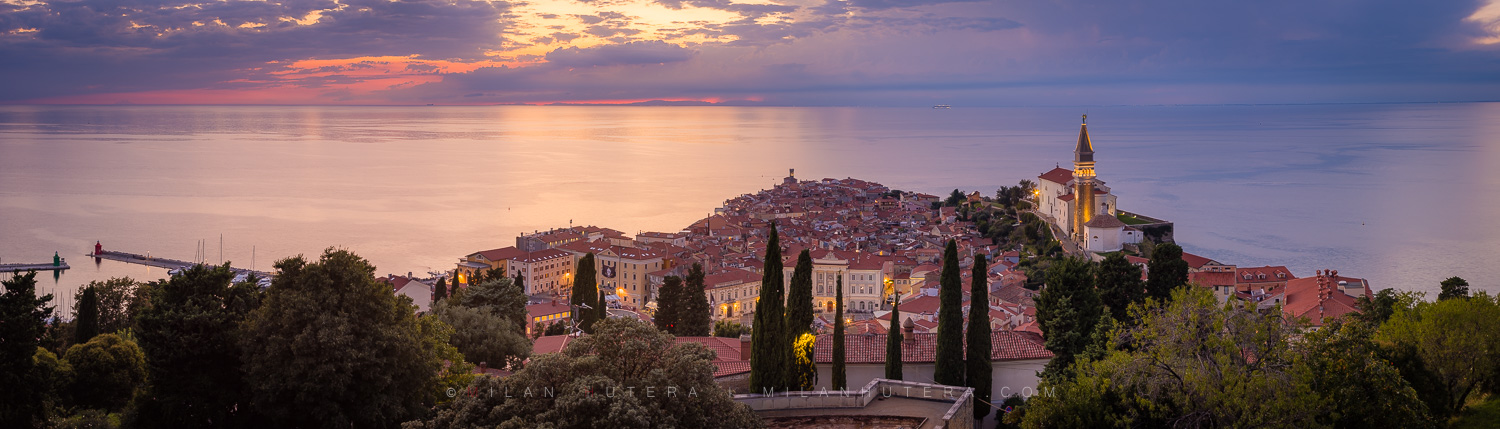Panoramic sunset view of Piran old town from the Piran Walls, Slovenia, featuring the St. George's Parish Church bell tower and the Adriatic Sea.