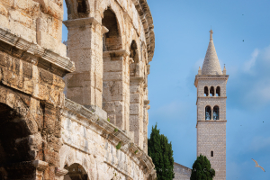 Roman amphitheater Pula Arena with ancient stone arches in the foreground and the white stone church tower of St. Anthony under a blue sky in Croatia.