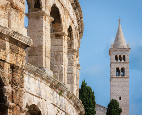 Roman amphitheater Pula Arena with ancient stone arches in the foreground and the white stone church tower of St. Anthony under a blue sky in Croatia.