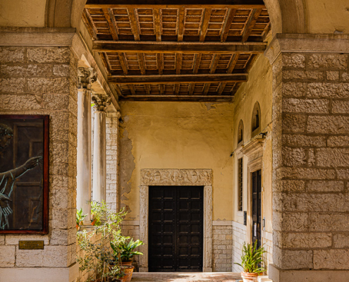 Side entrance archway of Pula Cathedral in Croatia with classical Roman columns, a weathered timber coffered ceiling, and potted Mediterranean plants