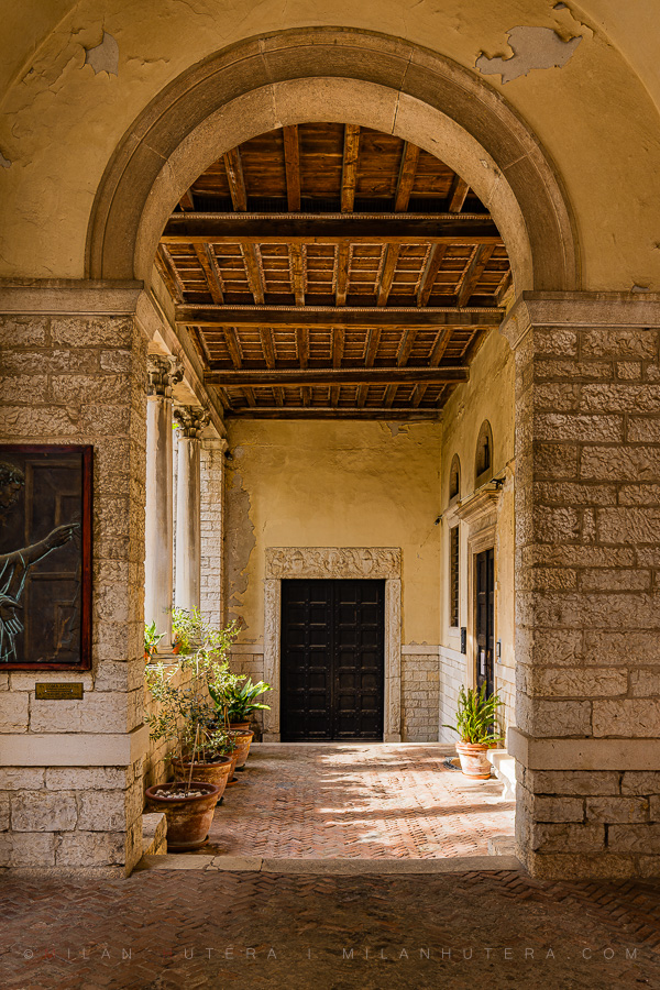 Side entrance archway of Pula Cathedral in Croatia with classical Roman columns, a weathered timber coffered ceiling, and potted Mediterranean plants