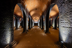 A perfectly symmetrical perspective looking down the main corridor of the 1894 Žlutý Kopec water reservoir, framed by massive brick columns and warm spotlighting.