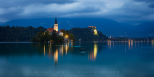 Evening panorama photography of Lake Bled at night, Slovenia, featuring the illuminated island church, Bled castle, and light trails from kayakers.