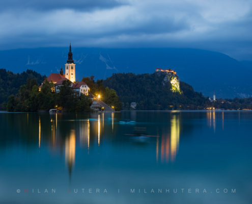 Evening panorama photography of Lake Bled at night, Slovenia, featuring the illuminated island church, Bled castle, and light trails from kayakers.