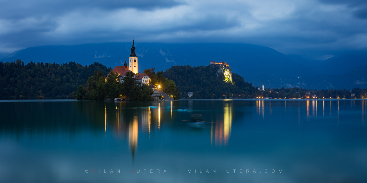 Evening panorama photography of Lake Bled at night, Slovenia, featuring the illuminated island church, Bled castle, and light trails from kayakers.
