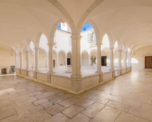 Wide-angle photography of the Minorite monastery courtyard in Piran, Slovenia, showing the beautiful Renaissance cloister, stone well, and vaulted arcades.