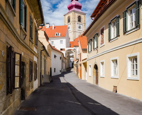 A scenic view looking up Aškerčeva ulica in Ptuj old town, leading toward the historic church tower under a blue sky.