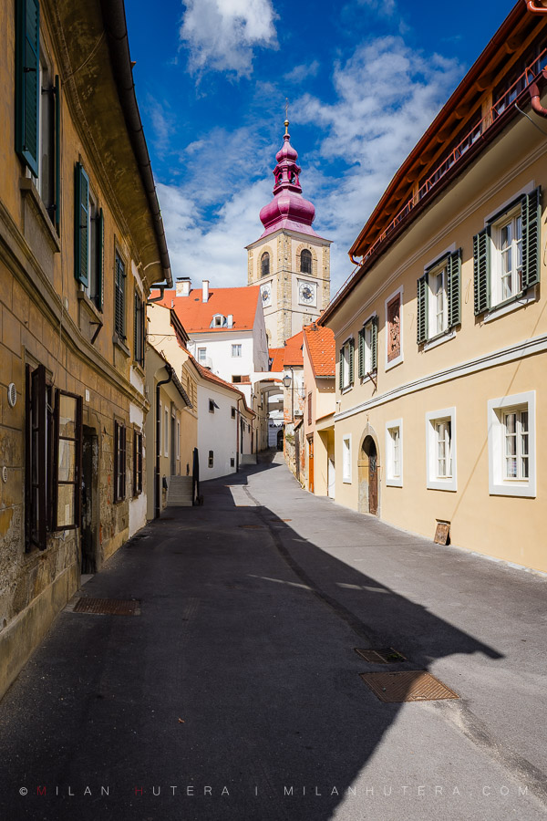 A scenic view looking up Aškerčeva ulica in Ptuj old town, leading toward the historic church tower under a blue sky.