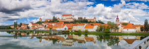 Ptuj panorama featuring the historic town and Ptuj Castle perfectly reflected in the calm waters of the Drava River under a cloudy blue sky.